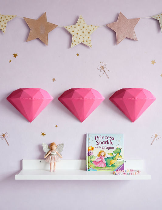 Decoración de pared para recámara de niña con tres diamantes geométricos en 3D en un solo color color rosa pastel, sobre una pared lila decorada con estrellas doradas y guirnalda de estrellas. Debajo, una repisa blanca con una muñeca de hada, un libro infantil de princesas y un brazalete de cuentas de colores.