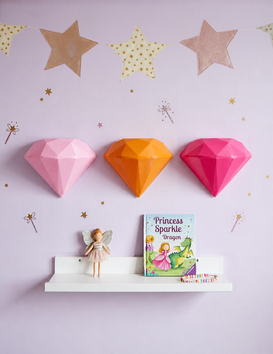 Decoración de pared para recámara de niña con tres diamantes geométricos en 3D en color rosa pastel, naranja y rosa mexicano, sobre una pared lila decorada con estrellas doradas y guirnalda de estrellas. Debajo, una repisa blanca con una muñeca de hada, un libro infantil de princesas y un brazalete de cuentas de colores.